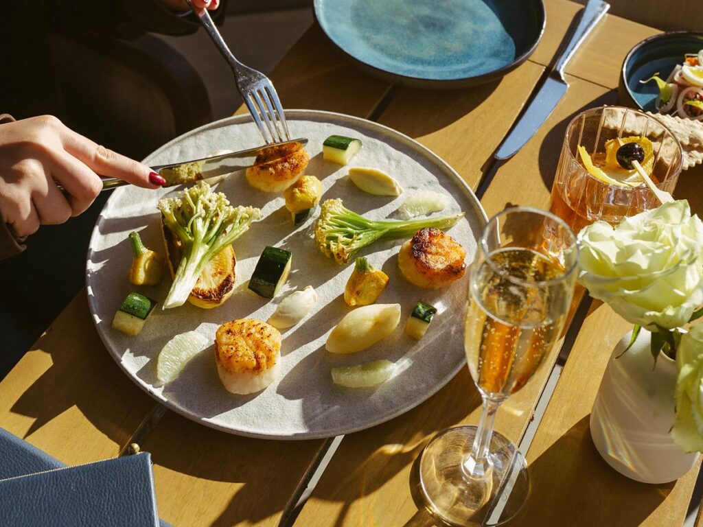 Seared scallops with seasonal vegetables served with champagne and a craft cocktail on a sunny terrace at L'Auberge Del Mar, luxury oceanfront dining in Del Mar, California